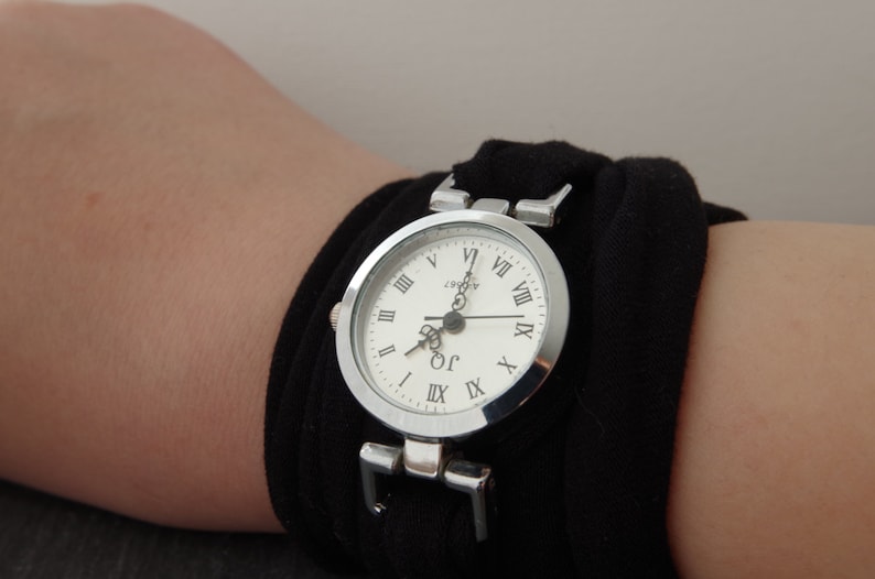 K&ouml;nnte beinhalten: Eine silberne Armbanduhr mit r&ouml;mischen Ziffern auf einem wei&szlig;en Zifferblatt. Die Uhr wird an einem schwarzen Stoffarmband getragen.