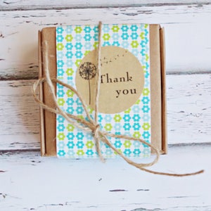 Puede incluir: Una peque&ntilde;a caja marr&oacute;n envuelta en papel estampado azul y verde con una pegatina circular marr&oacute;n que dice "Gracias" y un gr&aacute;fico de diente de le&oacute;n.