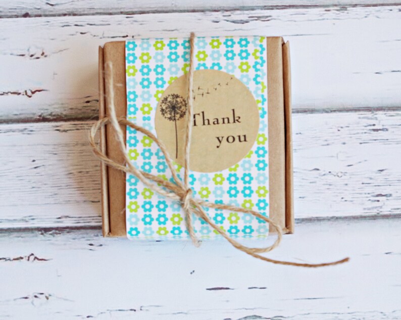 Puede incluir: Una peque&ntilde;a caja marr&oacute;n envuelta en papel estampado azul y verde con una pegatina circular marr&oacute;n que dice "Gracias" y un gr&aacute;fico de diente de le&oacute;n.