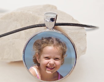 May include: A silver pendant necklace with a round glass charm featuring a smiling girl with blonde hair. The girl is wearing a pink swimsuit and is in front of a blue background.