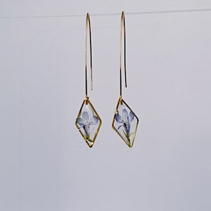 Könnte beinhalten: Ein Paar goldene Ohrringe mit einem diamantförmigen Anhänger mit einer gepressten blauen Blume.