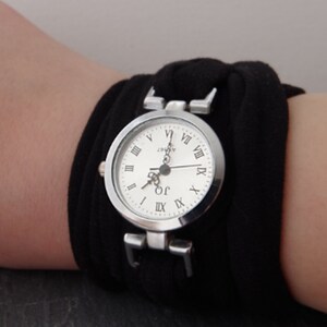 K&ouml;nnte beinhalten: Eine silberne Armbanduhr mit r&ouml;mischen Ziffern auf dem Zifferblatt und einem schwarzen Stoffband.