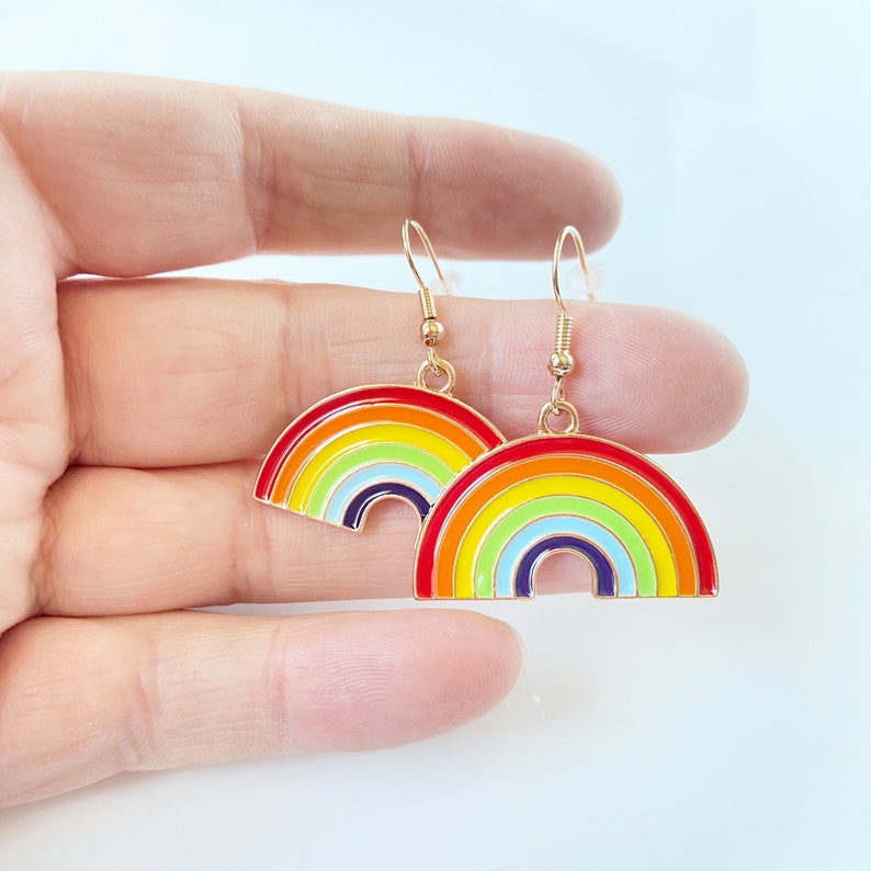 May include: A pair of gold dangle earrings with rainbow-coloured enamel. The earrings feature a rainbow arch design with red, orange, yellow, green, blue, indigo, and violet colours.