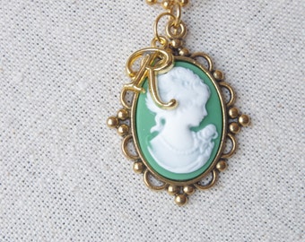 May include: A gold chain necklace with a green cameo pendant featuring a white silhouette of a woman's profile. The pendant has a gold letter 'R' charm attached to it.