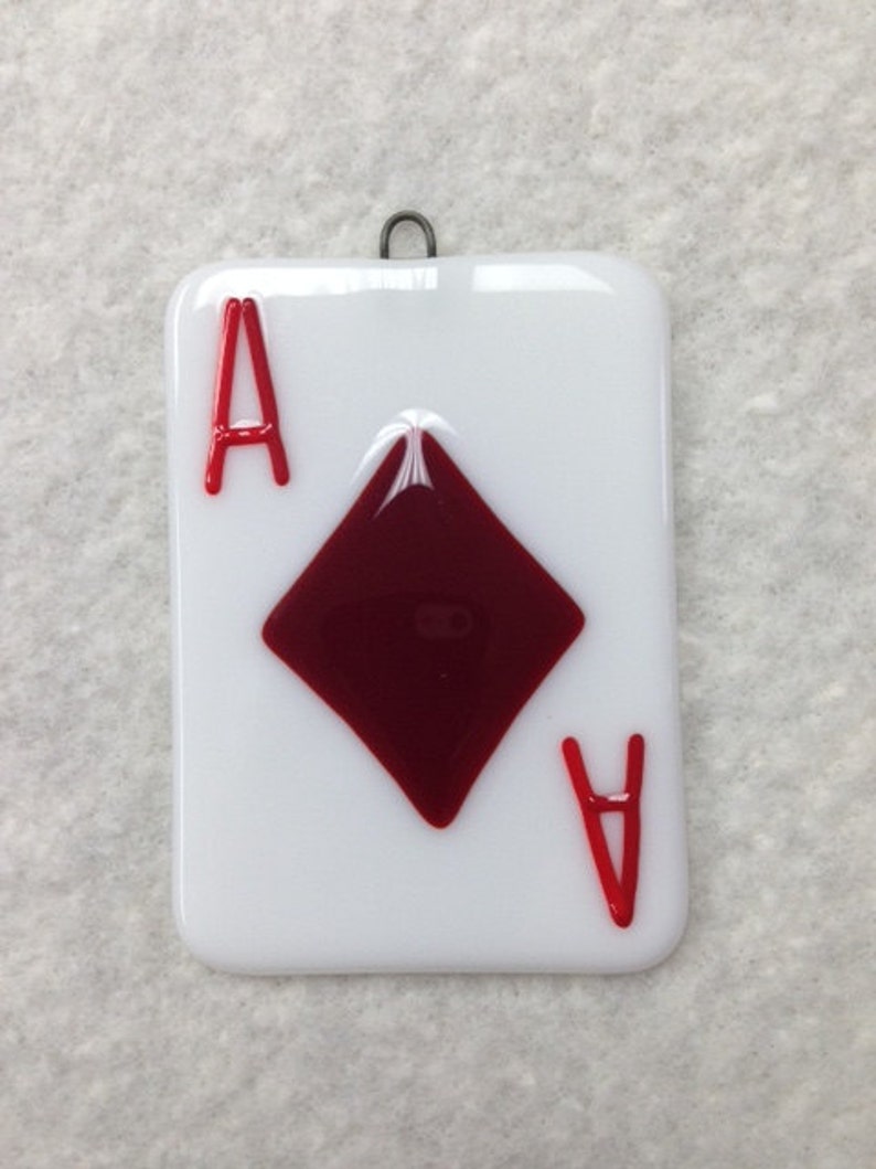 May include: A glass ornament shaped like a playing card, featuring the Ace of Diamonds. The card is white with a red diamond and red 'A's on each corner.