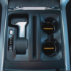May include: Interior car console with a gear shift, cup holders, and storage compartments. The cup holders have the word "TREMOR" in gold lettering. The gear shift is silver and black.