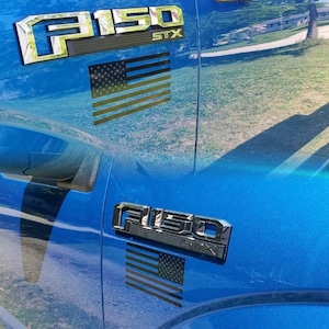 May include: A blue Ford F150 pickup truck with a black and white American flag decal on the side. The truck has a chrome "F150" emblem and "STX" lettering on the side.