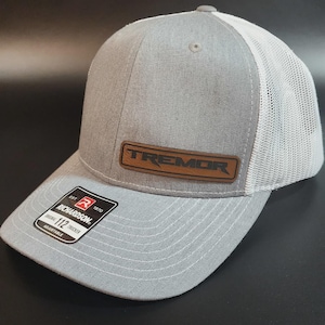 May include: A gray trucker hat with a white mesh back and a curved brim. The hat features a brown leather patch with the word "TREMOR" and a Richardson 112 Trucker label. The hat is adjustable.