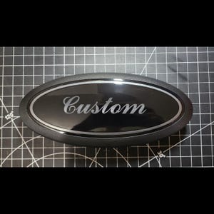 May include: A black oval emblem with the word "Custom" in a cursive font. The emblem has a silver border and is set against a black background. The item appears to be a car accessory.