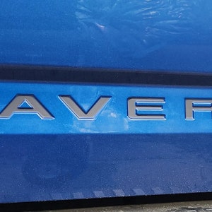 May include: A close-up of the back of a blue car with the word "RAVER" in black lettering on the tailgate.