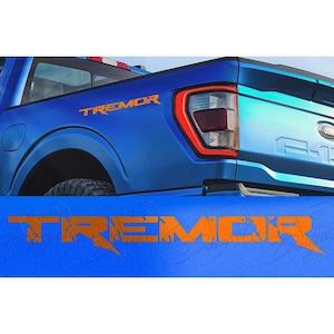May include: A blue Ford F-150 truck with orange "TREMOR" decals. The truck has a black tire and a red taillight. The word "TREMOR" is also displayed on a blue surface in the foreground.
