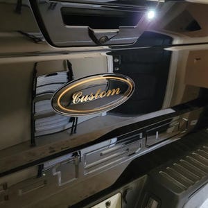 May include: Black truck tailgate with a custom gold and black oval emblem that reads "Custom". The Ford logo is visible below the emblem. The tailgate is glossy and reflects the surrounding environment.