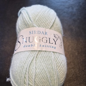 Op de afbeelding: Een bol lichtgroen Sirdar Snuggly dubbel breigaren. Het etiket is wit met de merknaam en producttype in donkerblauw. Het garen is strak opgerold en geschikt voor breiwerk.