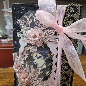 Könnte beinhalten: Ein dekoratives Journal mit einem schwarzen Einband, verziert mit rosa Spitze, floralen Verzierungen und einem rosa Band. Das Journal zeigt eine Illustration einer Frau mit floralem Kopfschmuck.