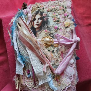 May include: A decorative journal featuring a woman's portrait, embellished with pink lace, ribbons, and floral accents. A gold clasp and pearl brooch are visible. The edges are decorated with colourful fabric tassels. The journal is a unique, handcrafted item.