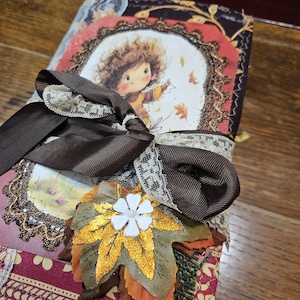 May include: A decorative journal with a fall theme. The cover features a whimsical illustration of a child and autumn leaves, framed with lace and ribbon. A brown ribbon and a leaf embellishment with a white flower accent the journal.