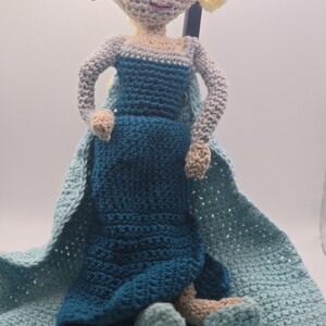 May include: A handmade crochet doll in the likeness of a princess, featuring a blue dress, light blue cape, and blonde hair. The doll is crafted with a textured stitch and has a soft, plush appearance. The doll is approximately 12 inches tall.