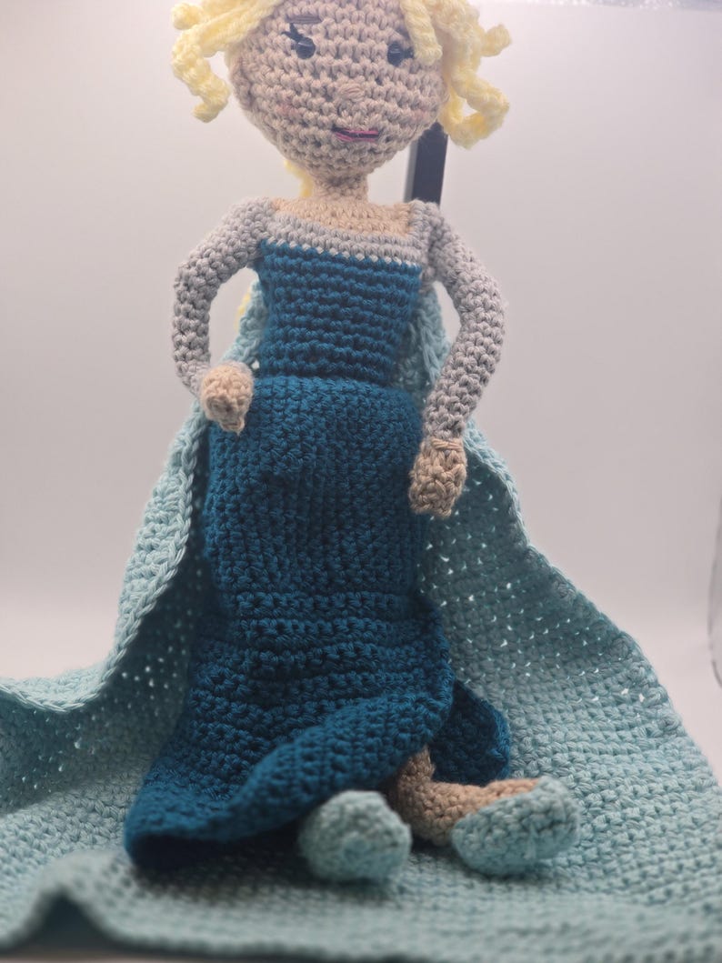 May include: A handmade crochet doll in the likeness of a princess, featuring a blue dress, light blue cape, and blonde hair. The doll is crafted with a textured stitch and has a soft, plush appearance. The doll is approximately 12 inches tall.