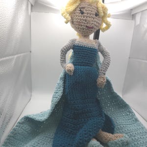 May include: Crocheted doll of a princess with blonde curly hair, wearing a blue dress and cape. The doll has gray arms and tan hands and feet. The dress and cape are shades of blue. The doll is handmade.