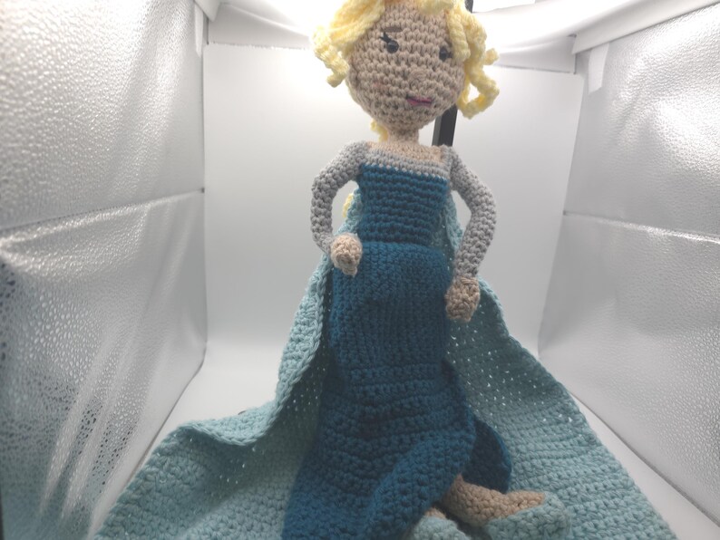 May include: Crocheted doll of a princess with blonde curly hair, wearing a blue dress and cape. The doll has gray arms and tan hands and feet. The dress and cape are shades of blue. The doll is handmade.