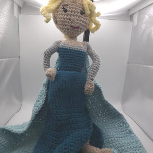 May include: A handmade crochet doll with blonde curly hair, wearing a blue dress and a light blue cape. The doll has a tan face and arms, and blue and tan shoes. The doll is sitting on a light blue crocheted blanket.