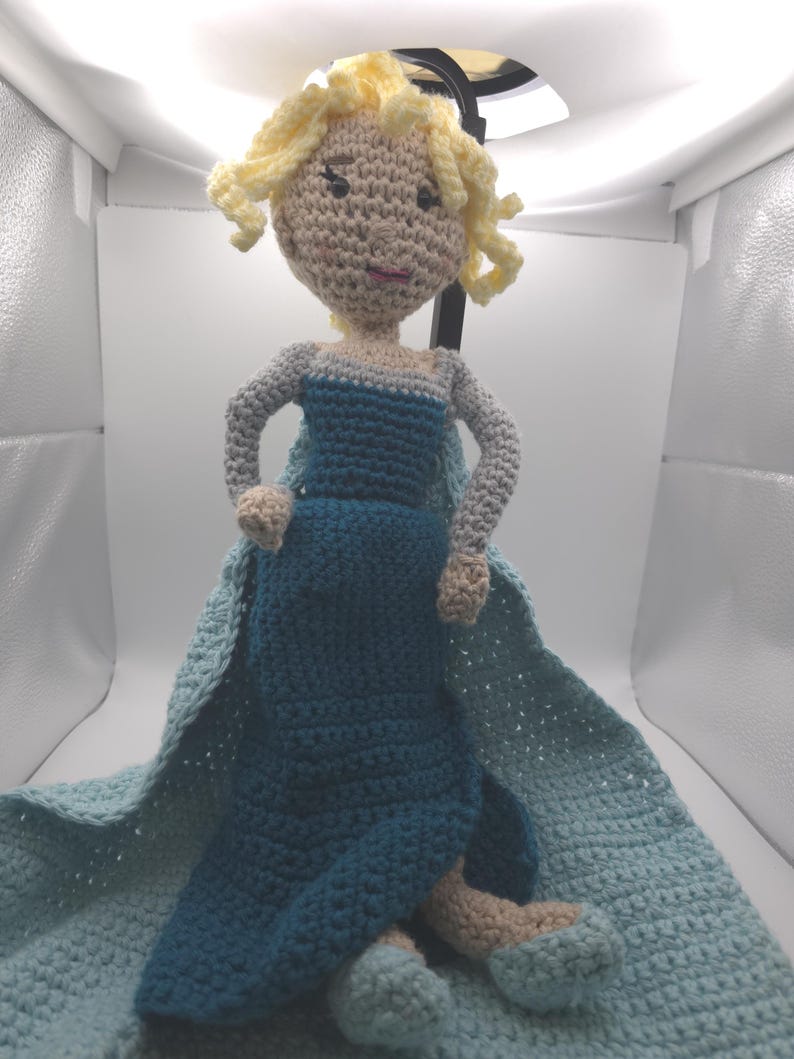 May include: A handmade crochet doll with blonde curly hair, wearing a blue dress and a light blue cape. The doll has a tan face and arms, and blue and tan shoes. The doll is sitting on a light blue crocheted blanket.