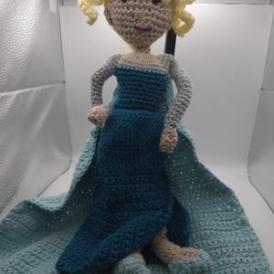 May include: A handmade crochet doll featuring a character with blonde curly hair, wearing a blue dress and cape. The doll is made with beige, blue, and light blue yarn. The doll is approximately 12 inches tall.