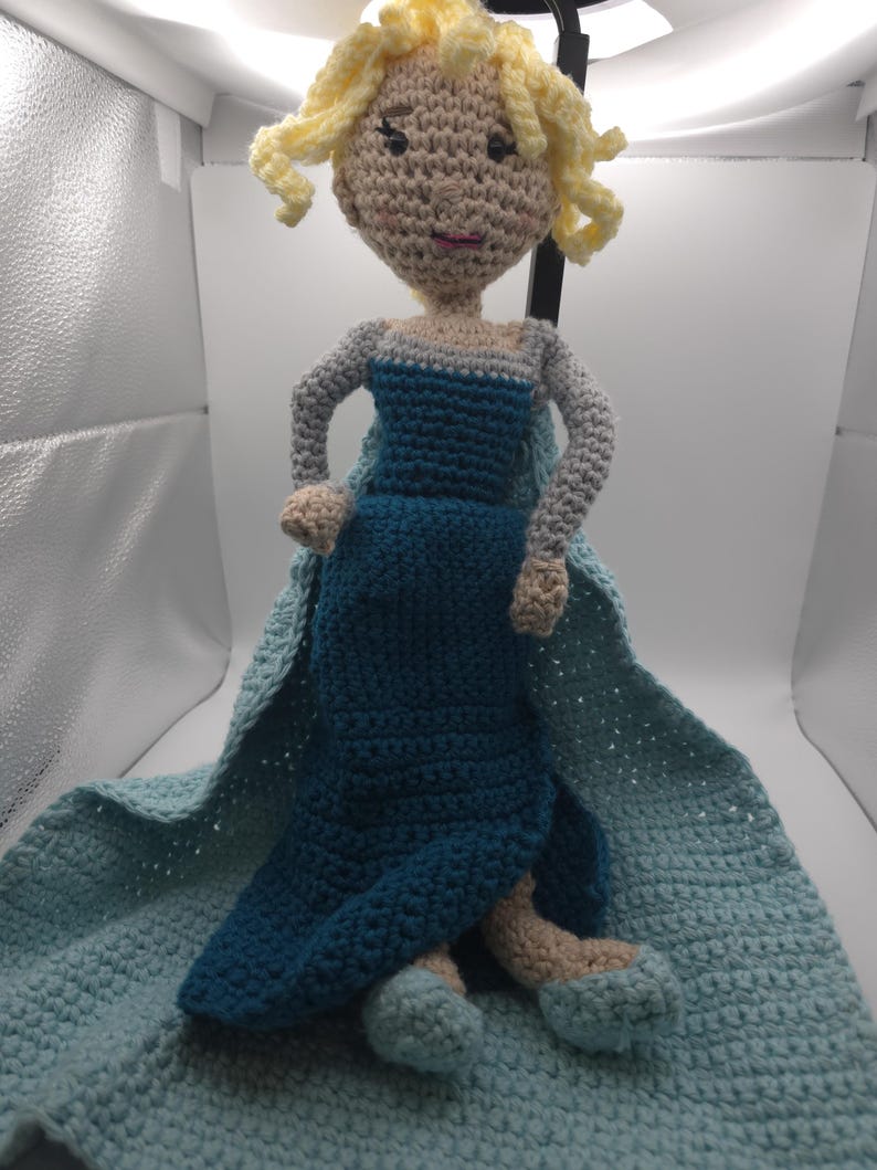 May include: A handmade crochet doll featuring a character with blonde curly hair, wearing a blue dress and cape. The doll is made with beige, blue, and light blue yarn. The doll is approximately 12 inches tall.