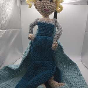 May include: A handmade crochet doll featuring a light skin tone, blonde curly hair, and a blue dress with a cape. The doll is sitting on a blue crocheted blanket. The doll's arms and legs are also crocheted.