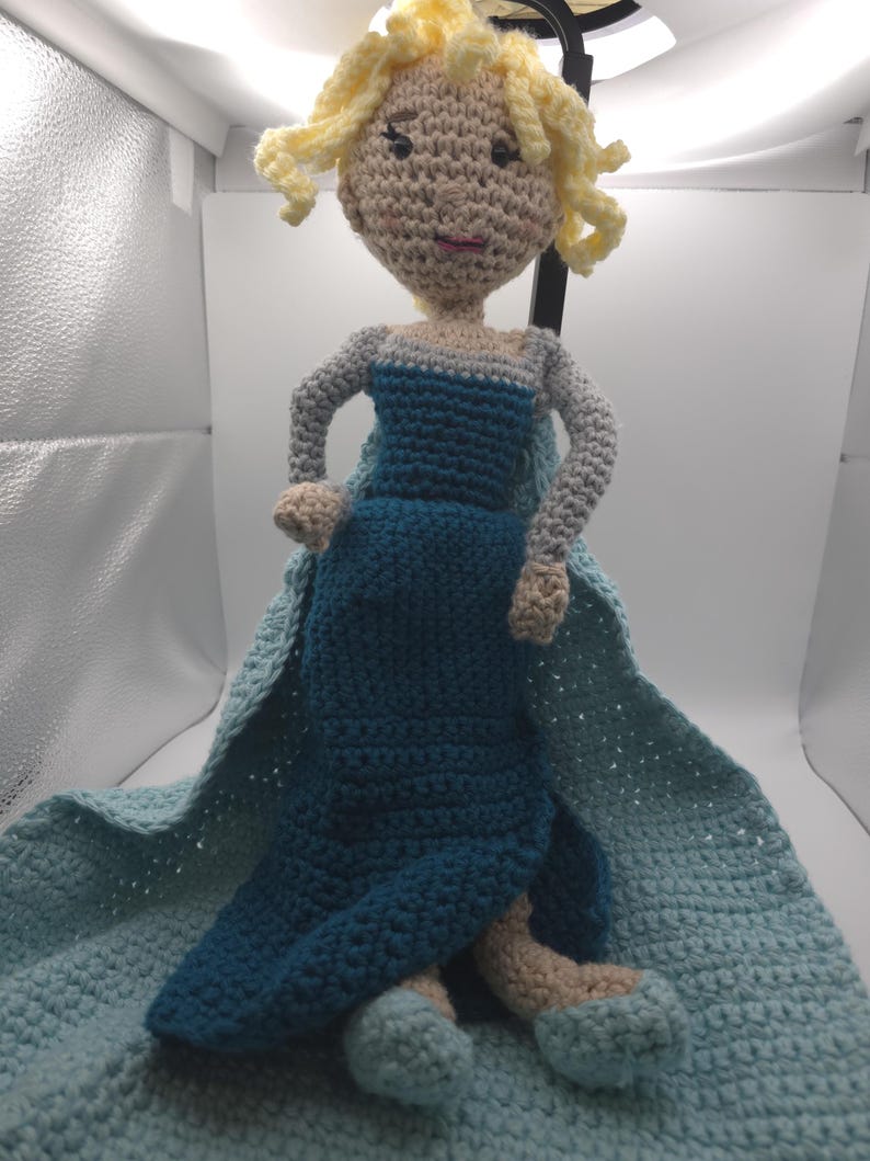 May include: A handmade crochet doll featuring a light skin tone, blonde curly hair, and a blue dress with a cape. The doll is sitting on a blue crocheted blanket. The doll's arms and legs are also crocheted.