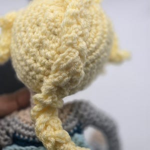 May include: Close-up of a crocheted doll's head with a light yellow yarn braid. The doll's head is a light yellow color, and the braid is intricately woven. The doll's clothing is visible in the background, with shades of blue and gray.