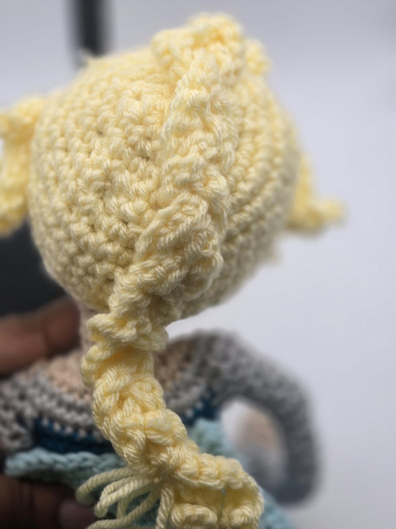 May include: Close-up of a crocheted doll's head with a light yellow yarn braid. The doll's head is a light yellow color, and the braid is intricately woven. The doll's clothing is visible in the background, with shades of blue and gray.