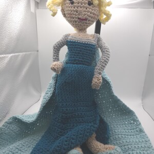 May include: A handmade crochet doll of a princess in a blue dress with a cape. The doll has blonde curly hair and is sitting on a light blue blanket. The doll's arms and legs are beige.