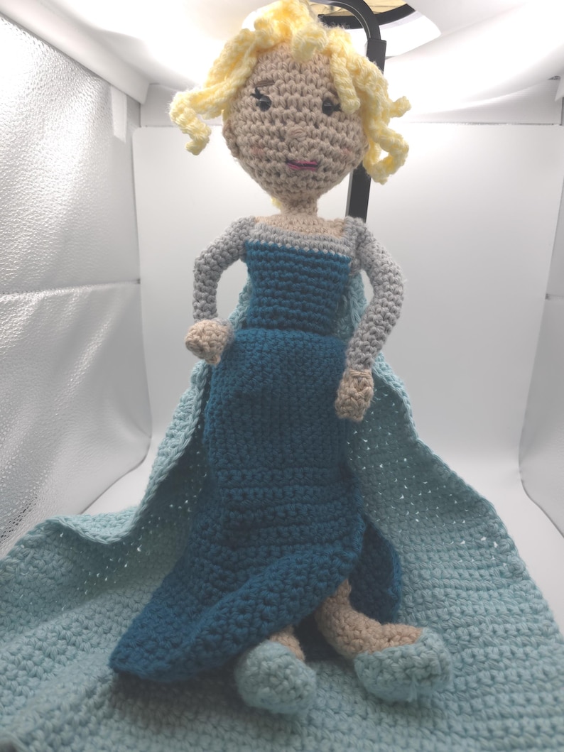 May include: A handmade crochet doll of a princess in a blue dress with a cape. The doll has blonde curly hair and is sitting on a light blue blanket. The doll's arms and legs are beige.