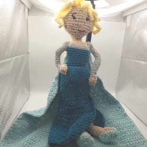 May include: A handmade crochet doll resembling a character from a popular animated film. The doll features a blue dress, a light blue cape, and blonde yarn hair. The doll is approximately 12 inches tall.