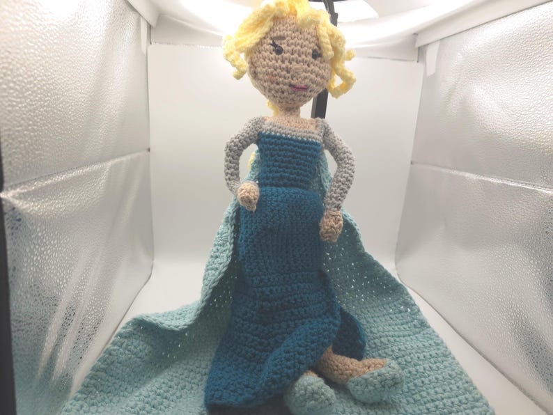 May include: A handmade crochet doll resembling a character from a popular animated film. The doll features a blue dress, a light blue cape, and blonde yarn hair. The doll is approximately 12 inches tall.