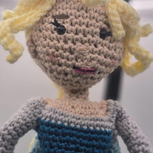 May include: Crocheted doll with blonde yarn hair styled in curls. The doll has a beige face with black eyes and pink lips. The doll is wearing a blue and gray dress.