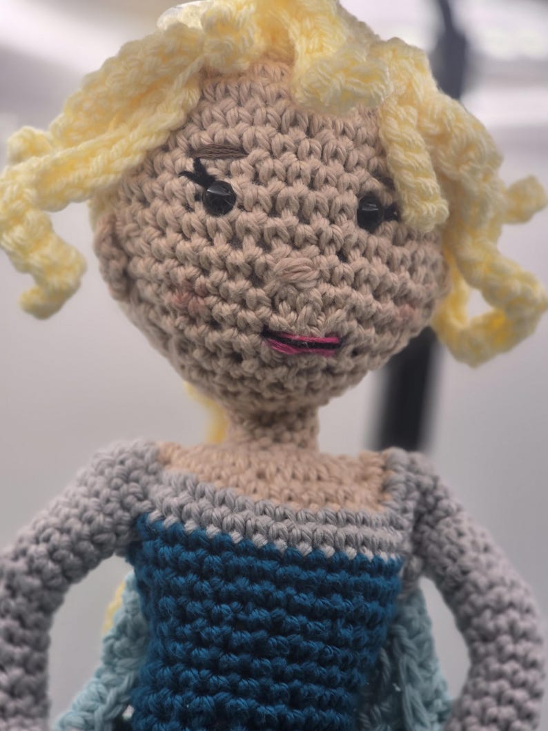 May include: Crocheted doll with blonde yarn hair styled in curls. The doll has a beige face with black eyes and pink lips. The doll is wearing a blue and gray dress.
