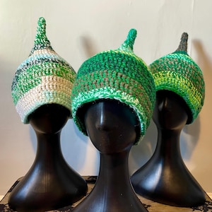 May include: Three green, white and brown crocheted hats with pointed tops. The hats are displayed on black mannequin heads.