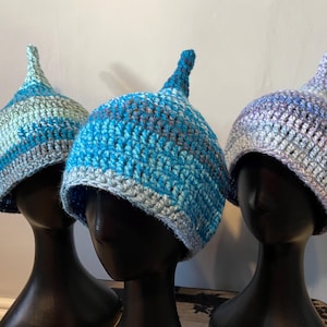 May include: Three crocheted hats with pointed tops in shades of blue, grey, and purple. The hats are displayed on mannequin heads.