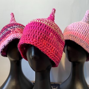 May include: Three pink and purple crocheted hats with pointed tops. The hats are displayed on black mannequins.
