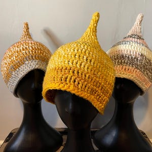 May include: Three crocheted hats with pointed tops in yellow, white and pink on black mannequin heads.