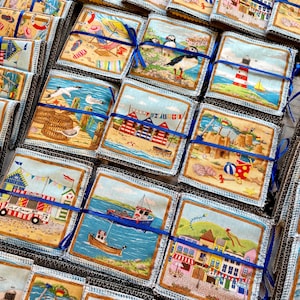 May include: A collection of colourful fabric squares featuring various seaside scenes, including beach huts, boats, and seabirds. Each square is tied with a blue ribbon and has a white border.