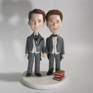 Könnte beinhalten: Zwei kleine Figuren von Bräutigamen in grauen Anzügen und weißen Hemden, die sich an den Händen halten. Der Bräutigam links trägt eine schwarze Fliege und ein schwarzes Stethoskop um den Hals. Der Bräutigam rechts trägt eine schwarze Fliege. Die Figuren stehen auf einem weißen Kuchensockel mit einem Stapel roter und goldener Bücher davor.
