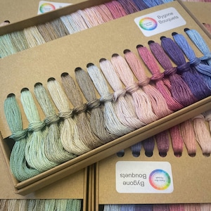 Könnte beinhalten: Eine Schachtel mit Bygone Bouquets Stickgarn in verschiedenen Farben, darunter Grün-, Beige-, Rosa- und Lilatöne. Die Fäden sind ordentlich gebündelt und in einer Pappschachtel angeordnet, wobei der Markenname auf einem Etikett zu sehen ist.