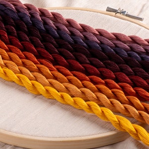 May include: A collection of embroidery floss spools in shades of orange, yellow, red, and purple. The floss is arranged in a gradient pattern, with the darkest shades at the top and the lightest shades at the bottom.