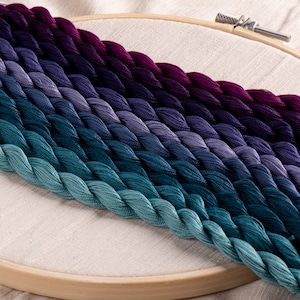 Pode incluir: Uma coleção de fio de bordado em tons de roxo, azul e verde, dispostos em um degradê em um bastidor de bordado de madeira.