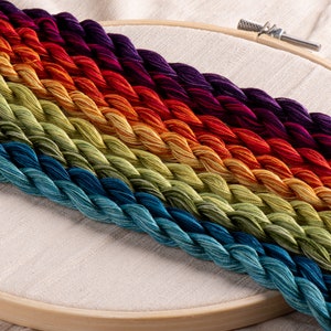 Peut inclure: Fils à broder multicolores disposés en arc-en-ciel dans un cercle à broder en bois. Les couleurs varient du bleu profond au violet, avec des nuances de vert, de jaune, d'orange et de rouge. Le tissu est beige clair.