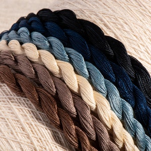 May include: A collection of embroidery floss spools in various shades of brown, beige, blue, and black. The spools are arranged in a fan shape, with the darkest colours at the top and the lightest colours at the bottom.