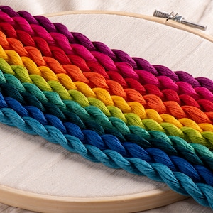 May include: A rainbow of embroidery floss in a wooden hoop. The floss is arranged in a gradient of colors, from deep blue to vibrant purple. The wooden hoop is partially visible, with the fabric stretched inside.